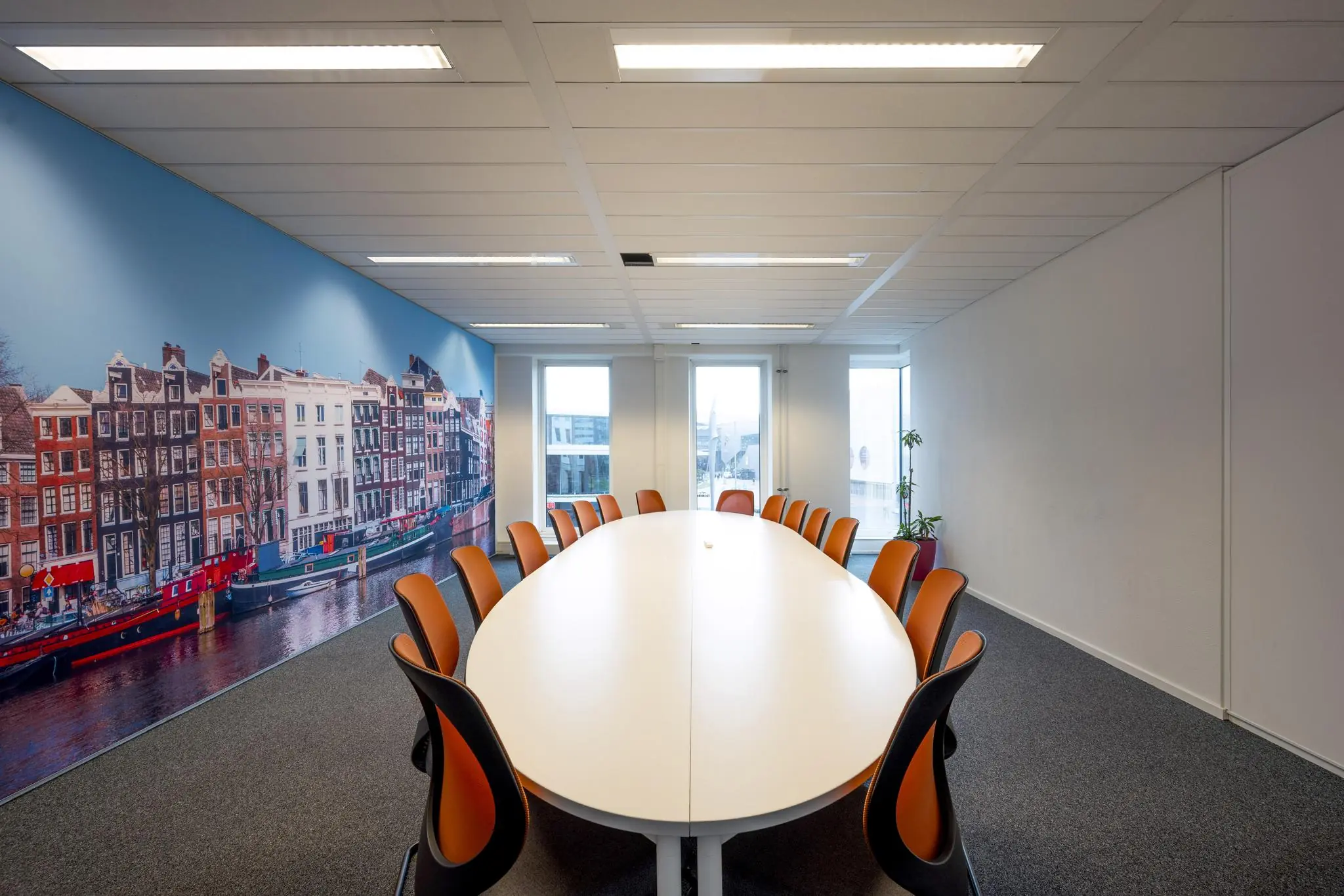 Vergaderruimte met ovale tafel, oranje stoelen en muurdecoratie van Amsterdamse grachtenpanden aan de Johan Cruijff Boulevard.